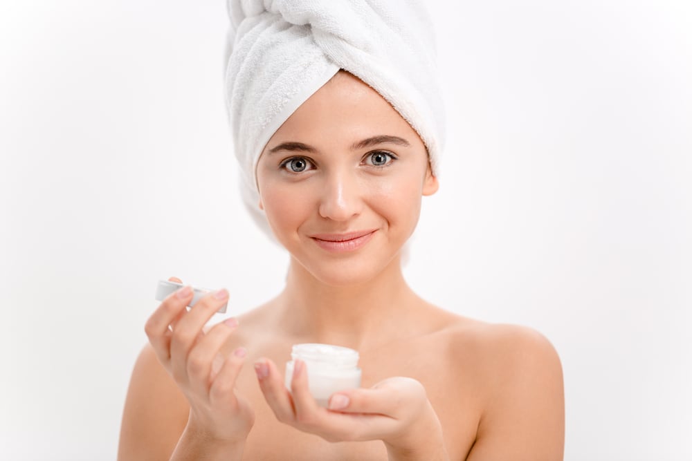 Beautiful young female with towel on her head using face cream over white background